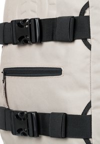 Bežový batoh vyrobený z odolného materiálu, s čiernymi plastovými sponami, zipsovou kapsou a detailmi prešívania.