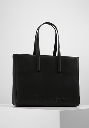Sort Calvin Klein tote taske med to lange håndtag, rektangulær form og subtilt præget logo på fronten, placeret på en hvid overflade.
