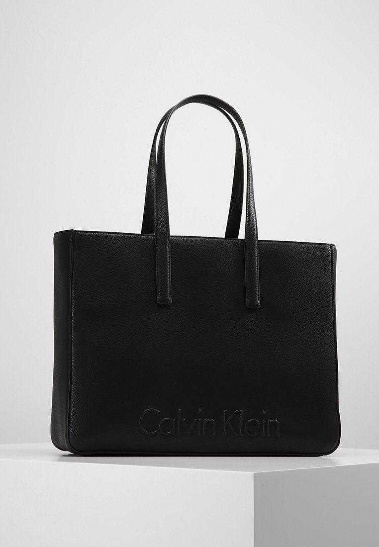 Sac cabas noir Calvin Klein avec deux longues anses, forme rectangulaire et logo subtilement embossé à l'avant, posé sur une surface blanche.