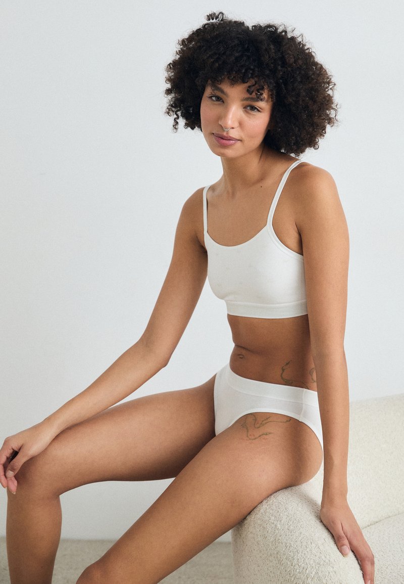 Donna con capelli ricci, vestita con biancheria intima bianca, seduta su una panchina di colore crema con texture, che guarda dolcemente verso la fotocamera.