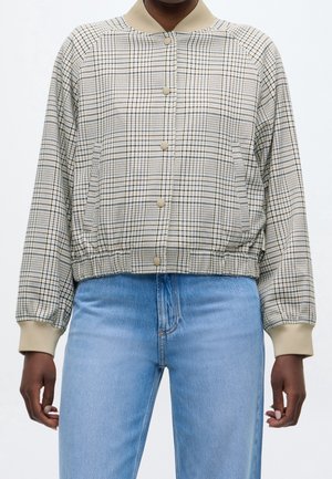 Personne portant une veste à boutons à carreaux beige et noir avec des poignets, un col et une ceinture beiges, assortie à un jean bleu clair.