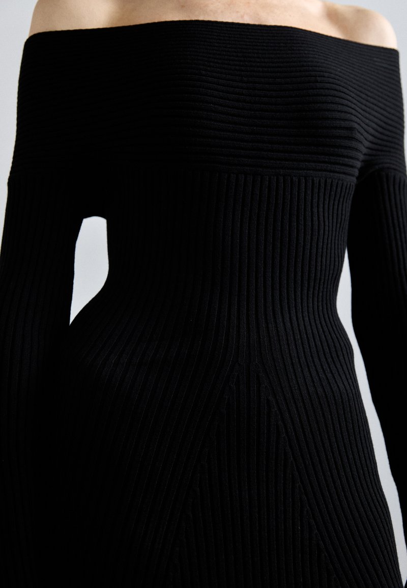 Robe noire côtelée à épaules dénudées avec des manches longues, présentant des lignes horizontales texturées et une silhouette ajustée.