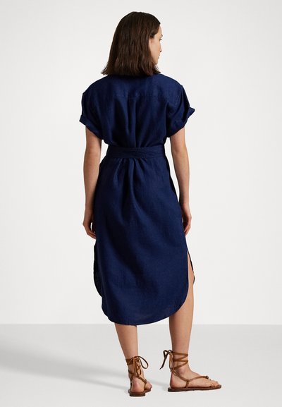 Jūras spēku krāsas lina kleita ar īsām piedurknēm, josta viduklī, sānu šķēlumi un noapaļots apakšmalas dizains. Kombinēta ar brūnām auklām sandālēm.