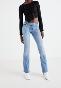 Haut long à manches longues en dentelle noire avec des détails découpés, assorti à un jean évasé bleu clair. Le mannequin porte des bottines blanches à talons.