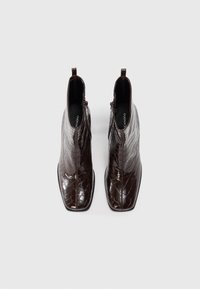 Par de botas de tobillo de charol marrón oscuro con estampado de cocodrilo, con punteras cuadradas y cremalleras laterales, vistas desde arriba sobre un fondo liso.