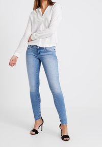 Blusa blanca de manga larga con cuello, combinada con jeans ajustados de color azul claro y sandalias negras de tacón. Tejido suave y diseño ajustado.