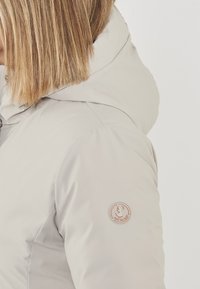 Giacca invernale grigio chiaro con cappuccio, texture liscia e una toppa circolare con logo sulla manica sinistra.