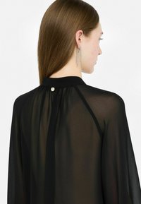 Doorzichtig zwart blouse met een hoge kraag, knoopsluiting op de rug en vloeiende mouwen, gemaakt van een lichte en transparante stof.