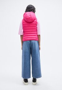 Ροζ μπουφάν χωρίς μανίκια με κουκούλα, συνδυασμένο με τζιν παντελόνι φαρδιάς γραμμής και μαύρα αθλητικά παπούτσια, φαινόμενο από πίσω σε λευκό φόντο.