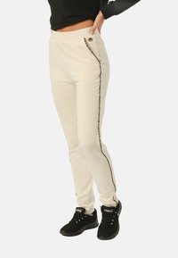 Pantalones jogger color crema con un corte ajustado, que cuentan con bolsillos laterales, acentos de estampado de leopardo negro y un logotipo dorado decorativo en la cintura.