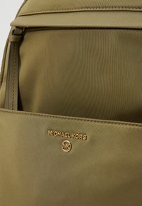 Maslinasto zelena najlonska ruksak s prednjim džepom na kojem se nalazi zlatni "Michael Kors" logo i kružni MK emblem. Glatka tekstura i elegantan dizajn.
