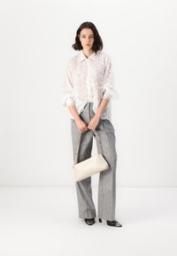 White, sheer, patterned blouse with wavy detailing, paired with gray plaid, wide-leg trousers. Model holds a beige, rectangular handbag.