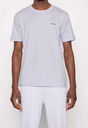 Homme portant un T-shirt à manches courtes lavande clair avec un petit logo "HUGO" sur la poitrine, associé à un pantalon de couleur claire.