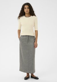 Ung kvinde med krøllet hår iført en cremet ribbet top, grå lang nederdel og sorte ballerinaer, står med hænderne i lommerne på hvid baggrund.