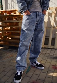 Ljusblå, löst sittande jeans i denim med en blekt struktur. I kombination med svart och vit rutiga sneakers. Träplattor i bakgrunden.