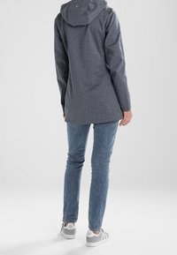 Šedý dlouhý kabát s kapucí, vyrobený z texturované látky; má rovný střih a minimalistický design; nošený přes modré džíny a tenisky.