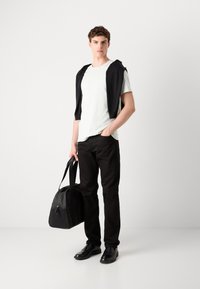 Mochila preta, t-shirt branca casual e calças escuras. Suéter de malha pendurado sobre os ombros. Sapatos pretos completam o visual.