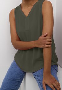 Haut sans manches couleur vert olive avec un col en V, présentant une texture subtile et un ourlet courbé. Associé à un jean ajusté bleu clair.