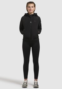Haut à zip noir avec une capuche doublée, des poignets côtelés et des poches, associé à des leggings noirs et des baskets noir et blanc.