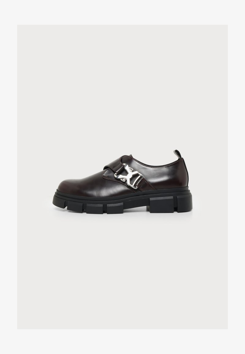 Mocassin en cuir marron foncé avec semelle noire épaisse et détail de boucle métallique sur fond blanc.