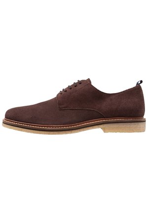 Scarpa da uomo in camoscio marrone con punta arrotondata, minimalista con lacci e suola in gomma chiara a contrasto. Presenta un piccolo tacco e dettagli di cucitura.