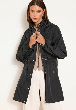 Naine, kes kannab musta pika vihmamantlit paeltest kinnitusdetailidega, beeži kardigani ja valgeid pükse, seisab ühevärvilise heleda tausta ees.