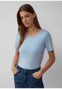 Lichtblauwe T-shirt met korte mouwen van een zachte stof, aansluitend ontwerp. Gecombineerd met donkerblauwe spijkerbroek. Model staat met handen in de zakken.