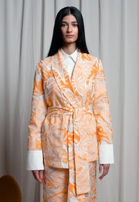 Blazer estampado em laranja e branco com calças combinando, apresentando uma cintura ajustável, colarinho de lapela e punhos brancos contrastantes.