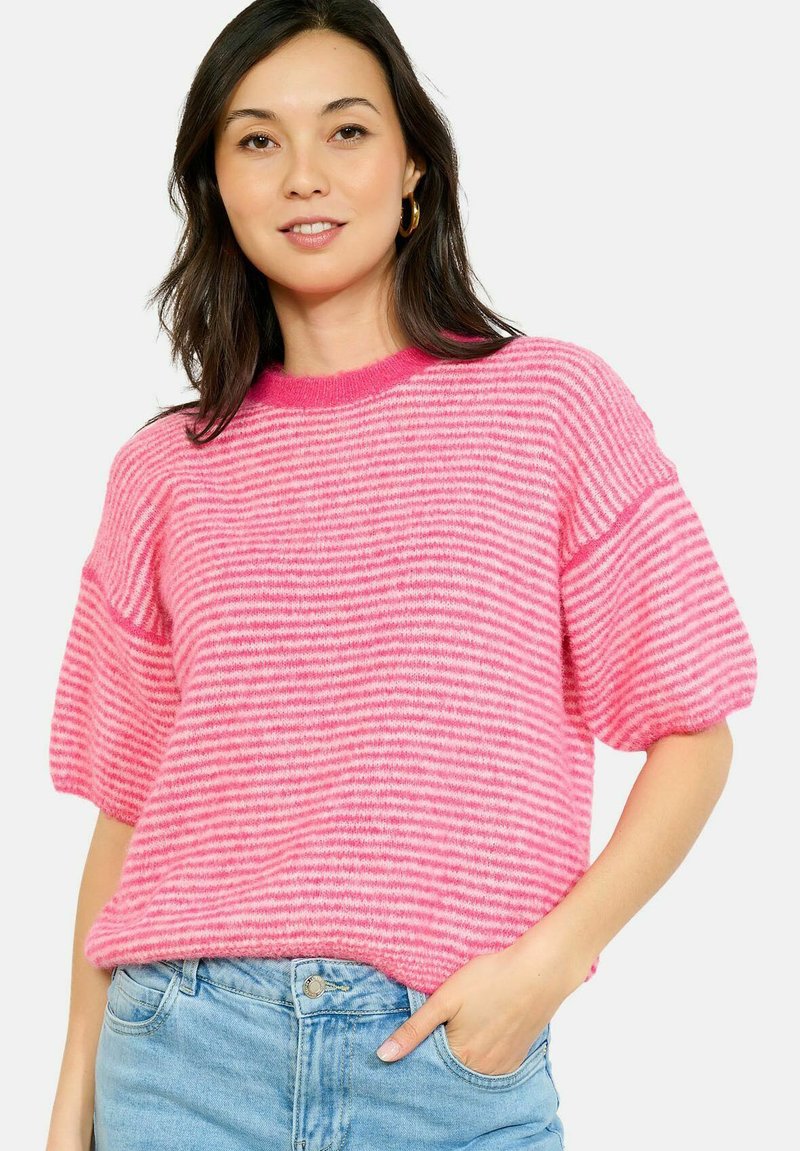 Young woman wearing pink striped fuzzy sweater with puffed sleeves and light blue jeans, standing with one hand in pocket.