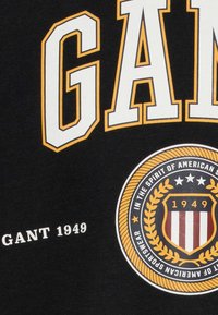 Czarny bawełniany rękawnik z napisem "GANT" w wyraźnym białym i żółtym kolorze, z okrągłym emblematem wykończonym na czerwono, biało i złoto.