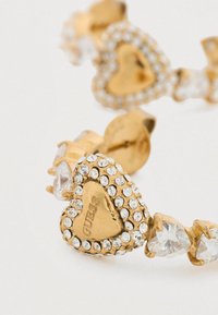 Βραχιόλι σε χρυσό τόνο σε σχήμα καρδιάς με διαφανή κρύσταλλα ρινοστούν, με υφή στην επιφάνεια και χαραγμένο λογότυπο "GUESS" στην καρδιά.