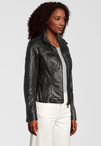 Zwarte leren jas met een rits aan de voorkant, textuurpanelen op de schouders, slim fit en ritszakken aan de zijkanten. Draagt over een grijze top.