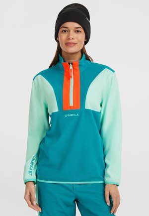 Teal fleecetrui met een halve rits kraag, oranje accent en wit logo. Kenmerkt zich door contrasterende mouwen en een "Polartec" detail op de manchetten.