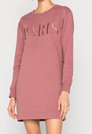 Kobieta ubrana w sukienkę dresową w kolorze pudrowego różu z długim rękawem i błyszczącym napisem "PARIS" na piersi.