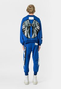 Conjunto de sudadera y pantalones azules con gráficos de alas en la parte posterior, con bordados texturizados y patrones en blanco y negro. Incluye detalles de etiqueta.