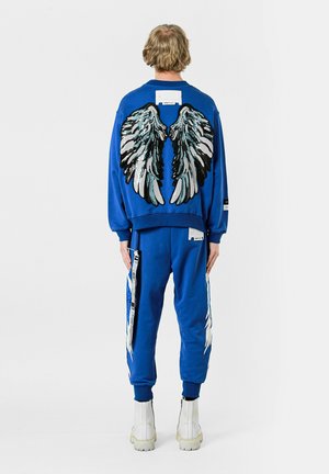 Conjunto de sudadera y pantalones azules con gráficos de alas en la parte posterior, con bordados texturizados y patrones en blanco y negro. Incluye detalles de etiqueta.