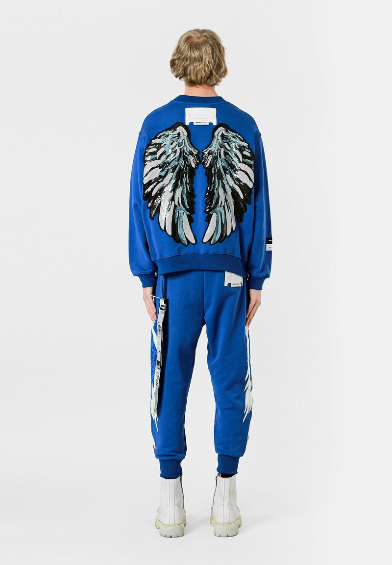 Conjunto de sudadera y pantalones azules con gráficos de alas en la parte posterior, con bordados texturizados y patrones en blanco y negro. Incluye detalles de etiqueta.