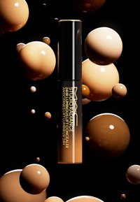 Il correttore luminosa 24 ore MAC Studio Radiance in un tubo cilindrico nero con testo dorato, circondato da sfere beige e marroni su uno sfondo nero.