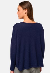 Femme avec des cheveux bruns raides portant un pull bleu marine ample et un pantalon gris, faisant face à la gauche avec le dos principalement visible.