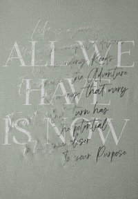 Miękka zielona tkanina z teksturowanym srebrnym i białym tekstem. Tekst różni się stylami czcionek, przekazując motywacyjne hasła i wzory.