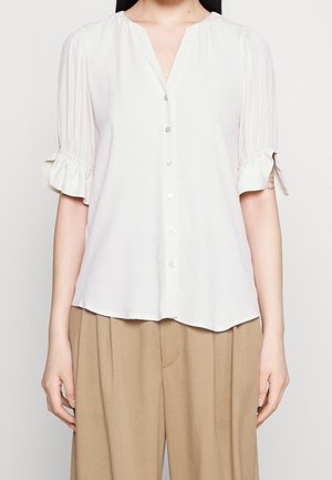 Kobieta w białej koszuli zapinanej na guziki z falbankami na krótkich rękawach i beżowych plisowanych spodniach o szerokich nogawkach na białym tle.