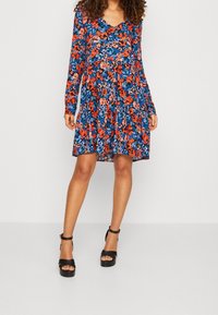 Květinové šaty s dlouhými rukávy, s námořnickým pozadím a jasnými červenými a modrými květy. Volný, ke kolenům sahající střih. Černé sandály na platformě.