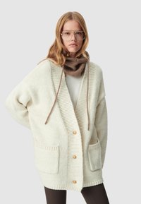 Cardigan a maglia crema con una vestibilità morbida, caratterizzato da una texture a coste, due tasche frontali e bottoni dorati. Accompagnato da una sciarpa marrone per il collo.