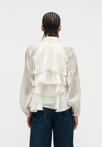 Άτομο με πλεκτά μαλλιά που φοράει διάφανη, κρεμ μπλούζα με βολάν και σκούρο μπλε τζιν παντελόνι, στέκεται γυρισμένο με την πλάτη σε έναν απλό φόντο.