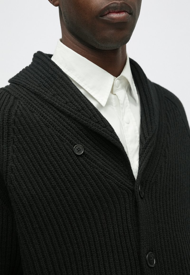 Cardigan nero lavorato a maglia con un design a coste testurizzate, dotato di colletto a scialle e due bottoni neri, indossato sopra una camicia bianca con colletto.