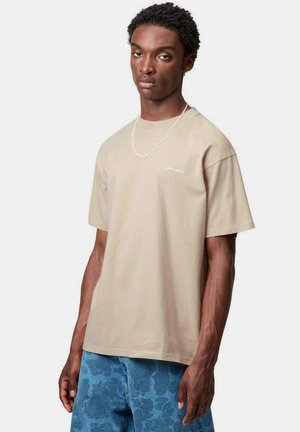 Camiseta de algodón beige de manga corta, con un pequeño logo blanco en el pecho. Llevada con unos jeans azules de estampado y un collar de cadena plateada.