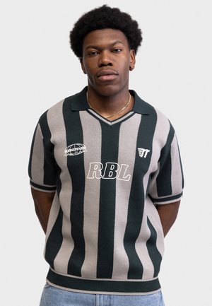 Młody mężczyzna stoi z rękami za plecami, ubrany w zielono-beżową koszulę w pionowe paski z krótkim rękawem, z napisem "RBL" i logo na piersi.