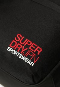 Tessuto nero con scritte ricamate in rosso e bianco che leggono "SUPER DRY JPN SPORTSWEAR" su un indumento sportivo.