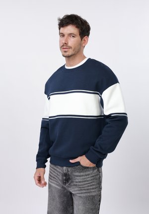 Muž s krátkymi tmavými vlasmi, ktorý má na sebe tmavomodrú a bielo pruhovanú mikinu a sivé rifle s vypraným efektom, stojí pred jednoduchým svetlým pozadím.