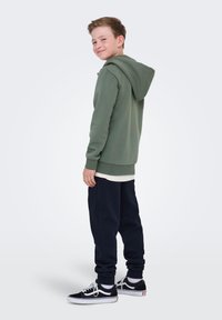 Niño sonriendo por encima del hombro, vestido con una sudadera verde, una camiseta blanca, pantalones deportivos azul marino y zapatillas negras, con un fondo claro y sencillo.